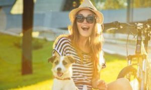 happy woman sits with little pet puppy beside bicycle in park in sunny sky