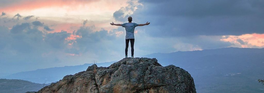 man stands on rock facing backward gratitude life in blue cloudy sky