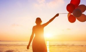 woman stands facing backward on beach holding balloons watching sunset