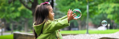 little girl stands in park playing with bubble