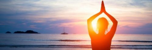 woman facing backward meditates on beach in blue sunny sky