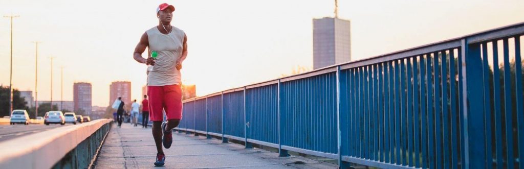 man holding water bottle wearing earphones jogging along footpath in city street