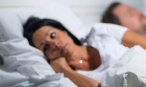 middle aged woman lying awake on bed while husband sleeping nicely