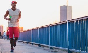 man holding water bottle wearing earphones jogging along footpath in city street