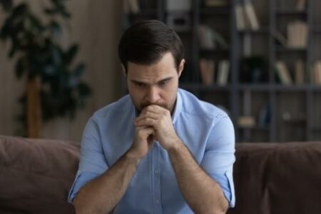 Confused unhappy young man lost in negative thoughts, considering personal problem solution, suffering from depression, feeling insecure or nervous waiting for important news, sitting on sofa at home.