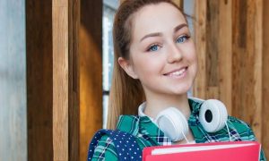 girl teenager wearing headphone stands in library holding red cover document smiling