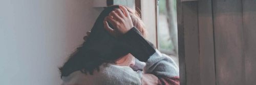 girl hands on head face downward sits beside window feeling sad