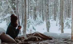 woman holding mug sits in house beside pillows watching snow