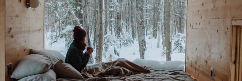 woman holding mug sits in house beside pillows watching snow