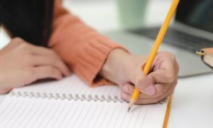 woman hand holding pencil writing on white notebook beside laptop