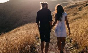 couple hand in hand facing backward walks in field talking in sunny sky