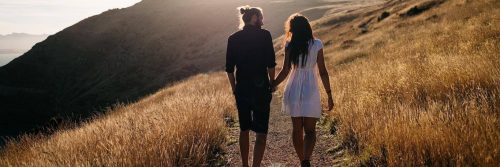 couple hand in hand facing backward walks in field talking in sunny sky