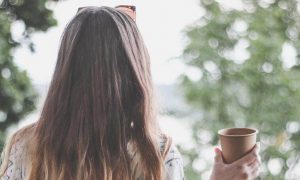 woman stands facing backward holding cup looking at trees lake