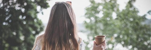 woman stands facing backward holding cup looking at trees lake