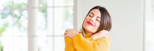 woman wearing yellow sweatshirt satisfied hugging herself in living room