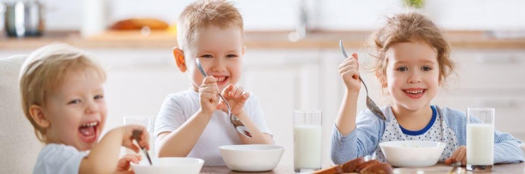 three kids happily sits eating healthy food drinking milk