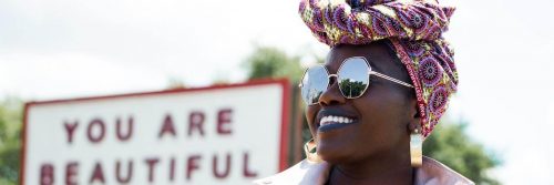 african woman wearing sunglasses stands beside street board happily smiling