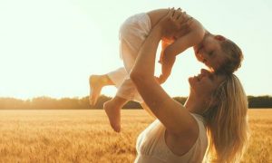 woman stands on field carrying little adorable son in sunny sky