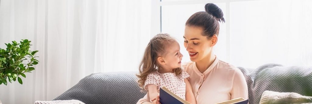 woman sits on couch smiling reading book for little adorable daughter in living room