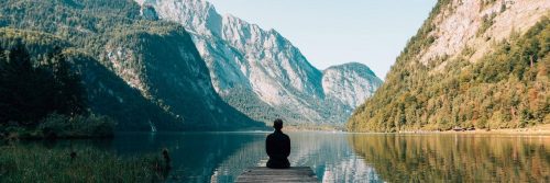 man sits on cliff facing backward looking at breakthrough mountain in blue clear sky