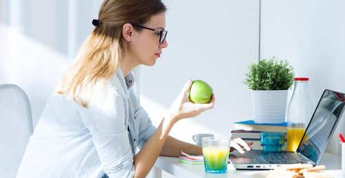 woman sits on chair holding green apple while working on laptop