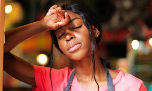 woman stands beside door hand on forehead eyes closed feeling tired