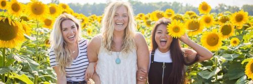 three women stand within sunflower garden happily smiling posing for photos