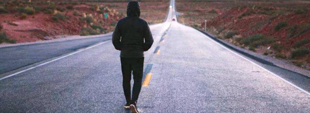 man wearing black hoodie facing backward walks alone on road between mountain