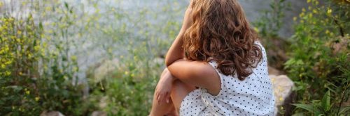 woman facing backward sits alone on rock beside green grass looking at river