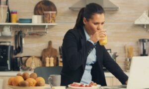 business woman stands in kitchen beside fruit dish drinks orange juice looks at laptop