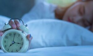 Girl hand on head holds white clock sleeps beside white pillows