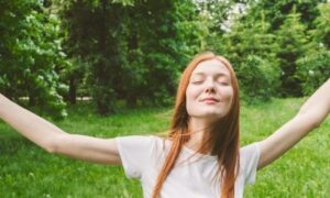 young woman raises hands eyes closed happily enjoys immerses into nature stands in forest