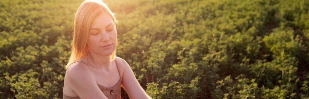 woman looks downward sits on green field in beautiful sunny sky