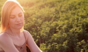 woman looks downward sits on green field in beautiful sunny sky