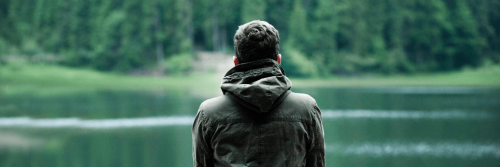 man stands alone facing backward looking at lake mountain