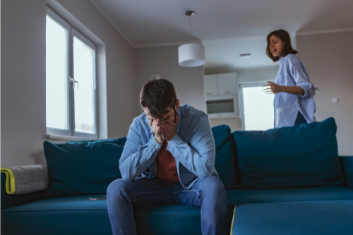 man sitting on blue couch face in hand crying  as woman yells angrily from across room