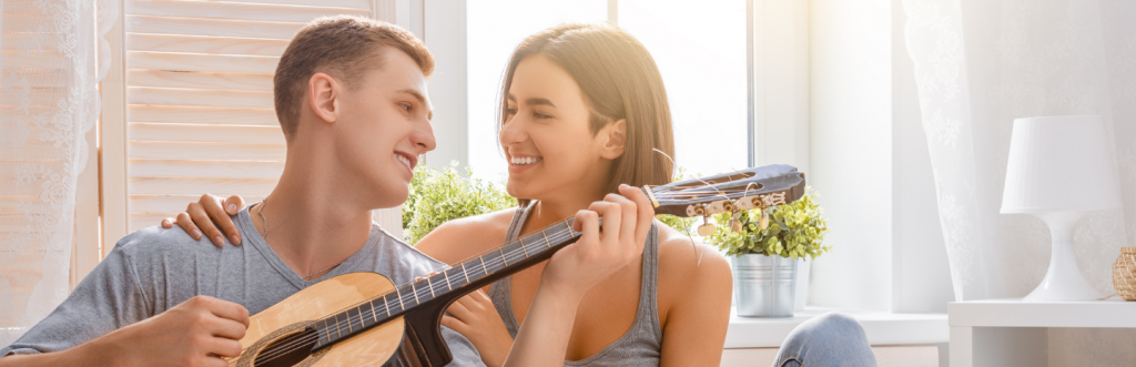 young man plays guitar happily smiles at girlfriend sitting next to