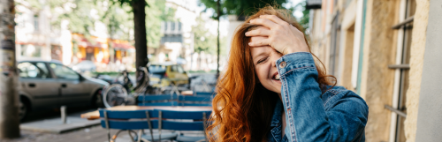 long curly brown hair woman with happy face hand on forehead laughs standing next to restaurant