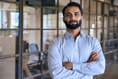 Confident happy successful ceo financial manager. Bearded indian businessman looking at camera standing in modern office with arms crossed. Handsome classy corporation owner. Business portrait.