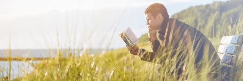 man sits on chair in field beside lake focusing on reading book in cloudy sky