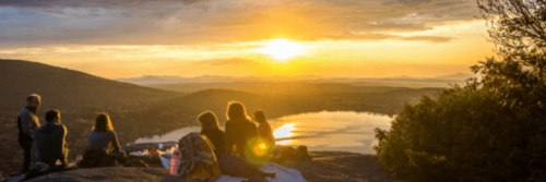 group of people sits on mountain top enjoying picnic watching sunset