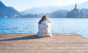 white hoodie girl sits alone facing blue ocean in quiet mountainous area