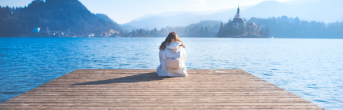 white hoodie girl sits alone facing blue ocean in quiet mountainous area