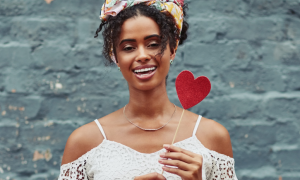 tanned skin girl happily smile carries red heart stick stands in blue stonewall background