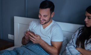 man sitting on bed chatting on mobile phone while girlfriend sits beside angrily unhappily starring at him