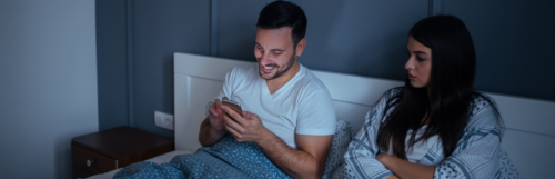 man sitting on bed chatting on mobile phone while girlfriend sits beside angrily unhappily starring at him