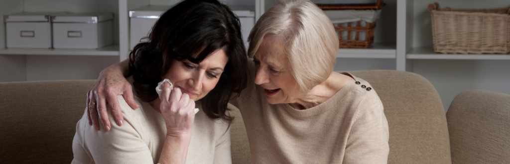 woman sits on couch in living room crying while elderly lady sitting beside comforting her