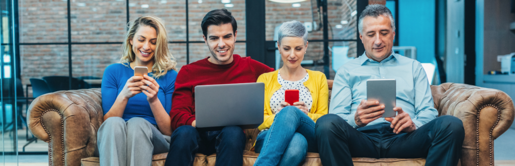 group of mixed gender from different generations sits on couch in office browsing social media on mobile phone laptop tablet