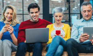 group of mixed gender from different generations sits on couch in office browsing social media on mobile phone laptop tablet