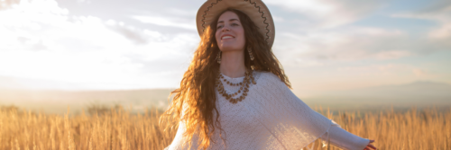 Beautiful young woman with a straw hat and a white poncho smiling and enjoying the sun in the middle of the grass field.